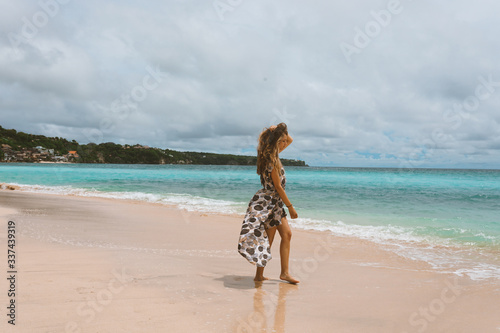 Beautiful and young girl in a dress posing on the shore of the ocean and sea with blue waves. Woman in a long dress model and fashion posing. Relax and relaxation on the island of Bali