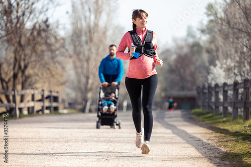 Pregnant young woman running in the park. In the background, her husband walking his son with the cart.