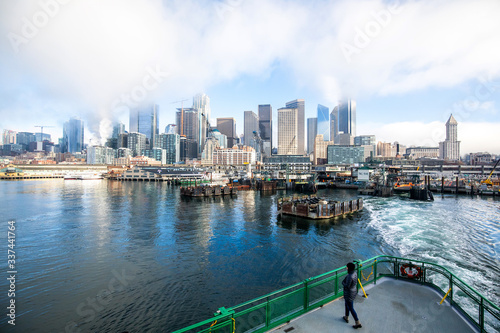 A person standing on the deck of a ferry boat in front of the Seattle waterfront on a cloudy morning. 