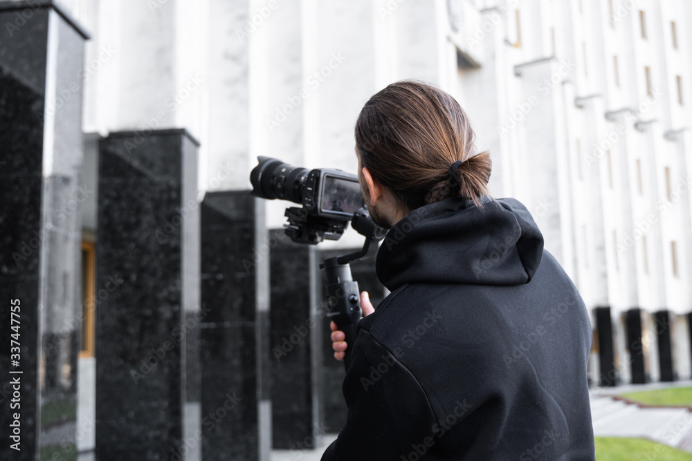 Young Professional videographer holding professional camera on 3-axis gimbal stabilizer. Pro equipment helps to make high quality video without shaking. Cameraman wearing black hoodie making a videos. Young Professional videographer holding professional camera on 3-axis gimbal stabilizer. Pro equipment helps to make high quality video without shaking. Cameraman wearing black hoodie making a videos.