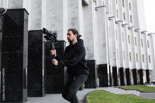 Young Professional videographer holding professional camera on 3-axis gimbal stabilizer. Pro equipment helps to make high quality video without shaking. Cameraman wearing black hoodie making a videos.