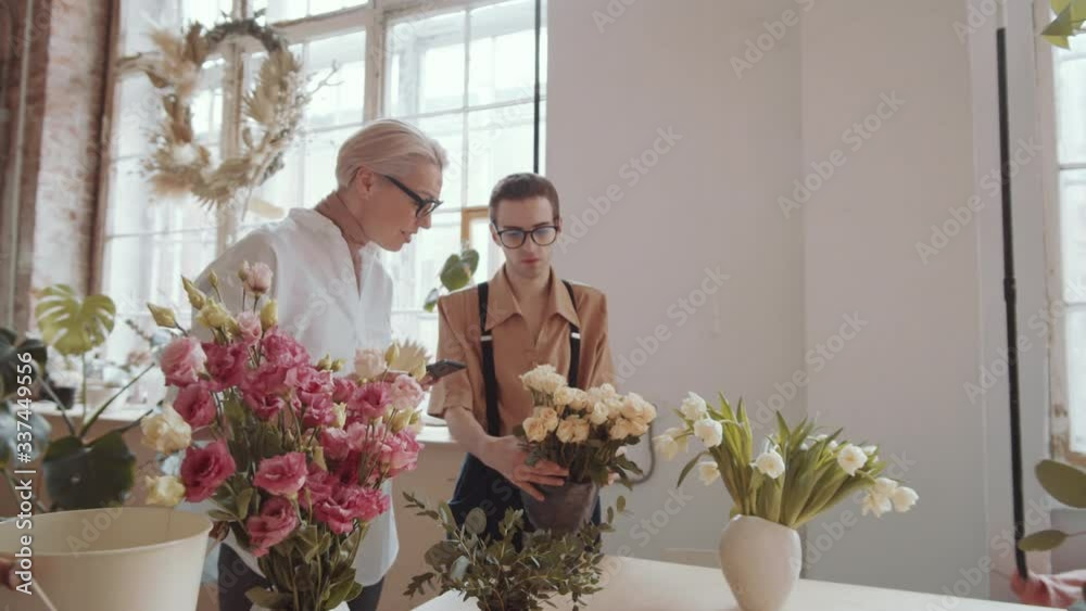 Zoom in shot of cheerful middle aged blonde woman typing on digital tablet and discussing roses with young male florist while working together in flower shop Zoom in shot of cheerful middle aged blonde woman typing on digital tablet and discussing roses with young male florist while working together in flower shop