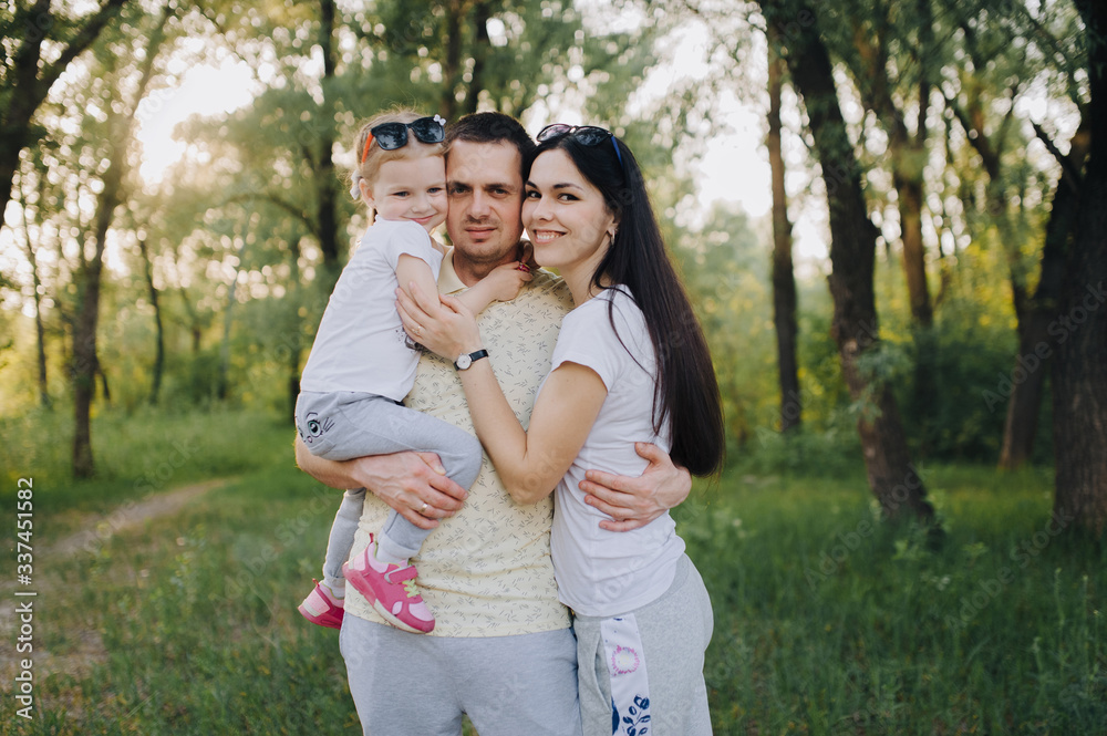 Portrait of a happy and smiling family in nature. A guy, a girl and a little daughter are hugging, standing in tracksuits in a green flowered garden and forest. Photography, concept.