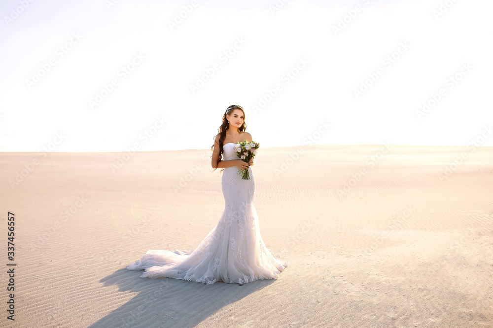 Bride in long wedding dress with bouquet stands on sand in desert. Backlight and pastel colors. Bride in long wedding dress with bouquet stands on sand in desert. Backlight and pastel colors.