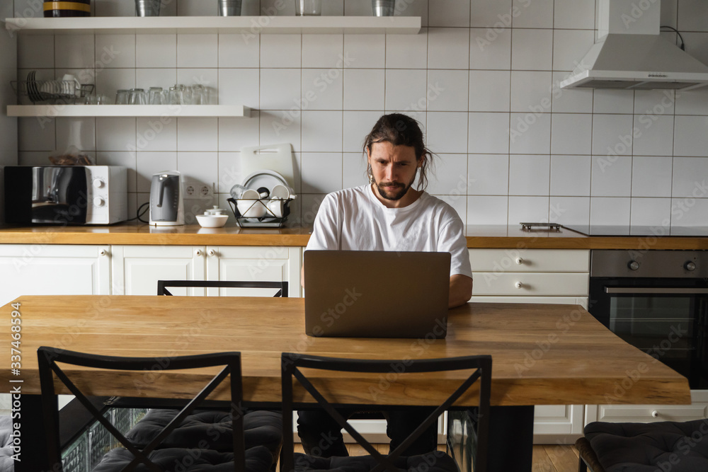 Freelancer working from home on the kitchen and using laptop. Bearded man working with a laptop and reading good news. Handsome successful self entrepreneur sitting and working at his modern home.