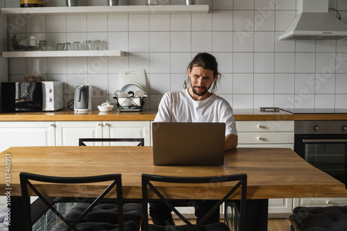 Freelancer working from home on the kitchen and using laptop. Bearded man working with a laptop and reading bad news. Handsome successful self entrepreneur sitting and working at his modern home.