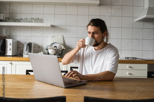 Freelancer working from home on the kitchen and using laptop. Handsome smiling man holding cup of coffee and looking at laptop. Successful self entrepreneur sitting with laptop and working. © Volodymyr