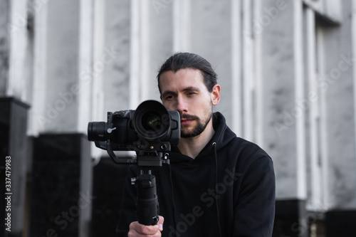 Young Professional videographer holding professional camera on 3-axis gimbal stabilizer. Pro equipment helps to make high quality video without shaking. Cameraman wearing black hoodie making a videos. © Volodymyr