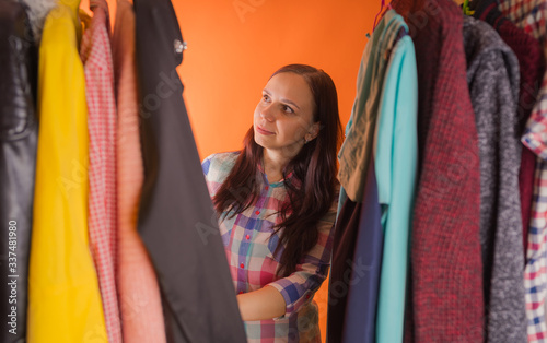 Young woman grimacing standing between clothes in wardrobe. Adult female choosing things in store.