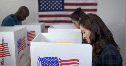 Side view MS of three people, two women and one man, Caucasian, Hispanic and African American, voting in booths at polling station. Large US flag on wall behind