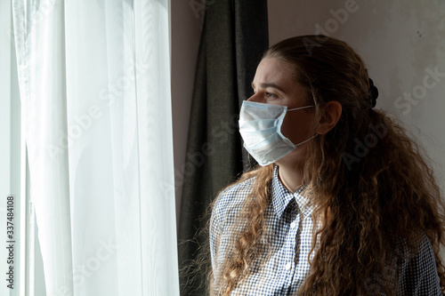 A young girl in a medical mask and gloves looks out the window while on self-isolation, quarantine at home. Home self-isolation in pandemic, flu epidemic, viral diseases, Covid-19.