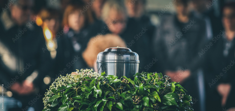 A metal urn with ashes of a dead person on a funeral, with people mourning in the background on a memorial service. Sad grieving moment at the end of a life. Last farewell to a person in an urn.