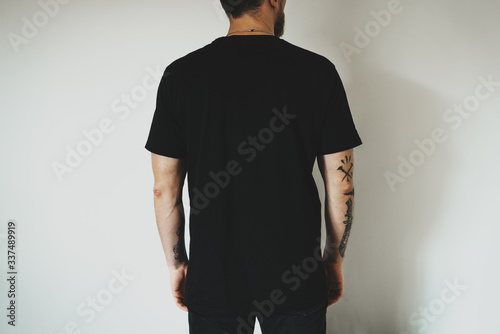 Young hipster wearing black jeans and blank black t-shirt with empty space for  logo, text or design. mock-up of t-shirt, white wall in the background. Back view.
