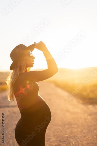 Beautiful caucasian pregnant woman with long hair in tight dress, sunglasses in sunlight on sunny desert road outdoors holds hat. Side view. New life, new horizons and opportunities concept