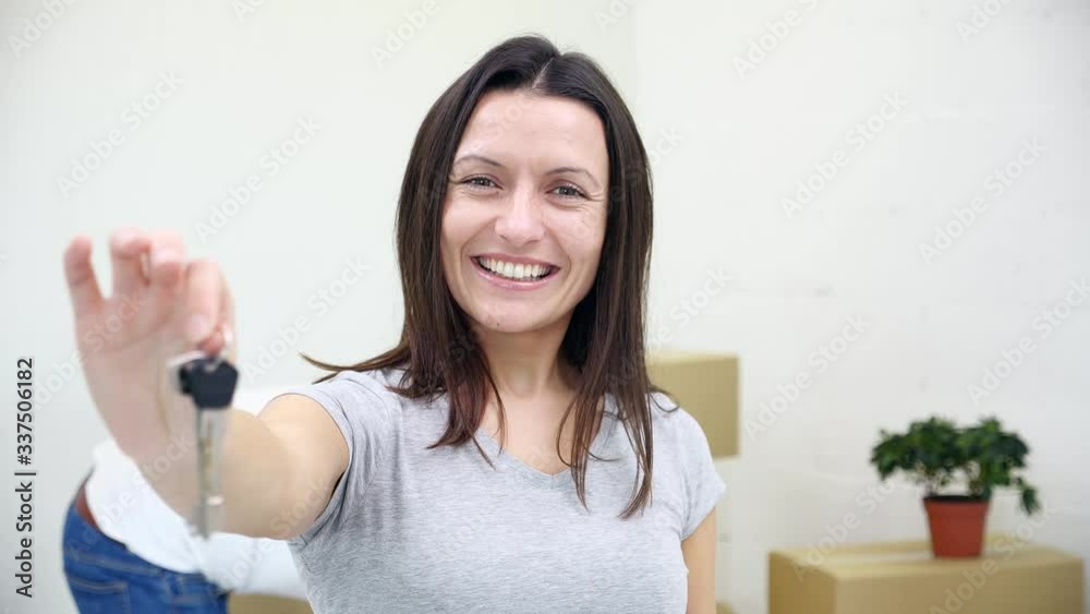 Toothy female apartment owner or renter showing keys to the camera whie her husband carrying boxes on the blurred background. Toothy female apartment owner or renter showing keys to the camera whie her husband carrying boxes on the blurred background.