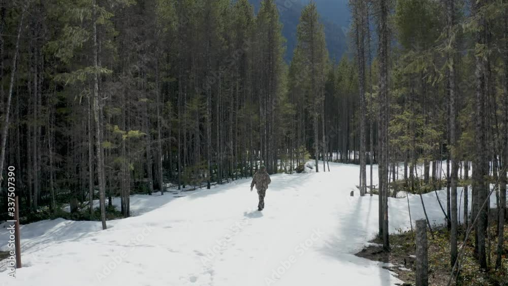 Elevated rear view of an outdoor man, wearing camouflage clothing and walking on a snowy trail through the forest. High Angle Shot Elevated rear view of an outdoor man, wearing camouflage clothing and walking on a snowy trail through the forest. High Angle Shot