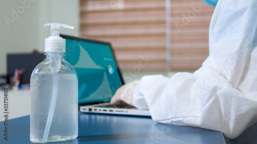 Close up view of a bottle of alcohol gel on a desk next to a doctor in a bioprotective suit working with his laptop in his office photo