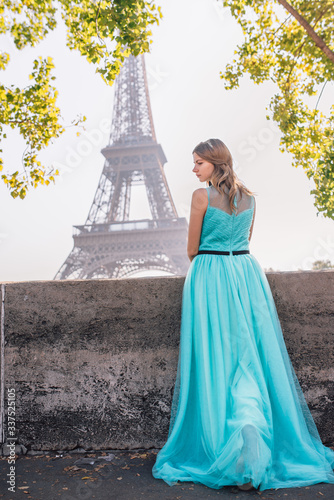 A young beautiful and elegant girl in a blue and green dress stands against the background of the Eiffel tower in Paris. © Aleksei Zakharov