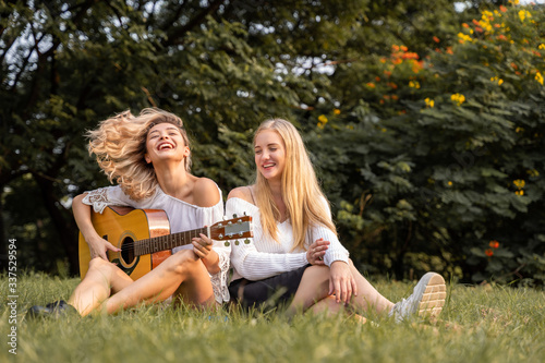 Portrait of caucasian young women sitting in the park outdoor and playing a guitar sing a song together with happiness