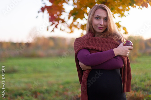Beautiful pregnant caucasian woman wrapped in warm sweater with apple in hand smiles and looks at camera. Outside on warm day. Nature soft focus backsides. Enjoying life and nature