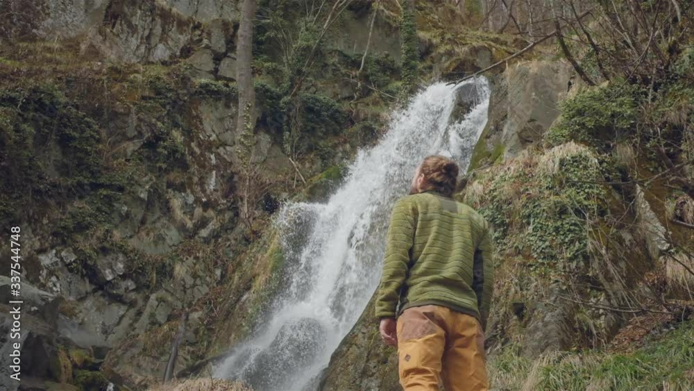 Young male traveler in enjoys a beautiful waterfall. Hiking in the mountains. The hiker runs to the waterfall, raises his hands up. Enjoys lifestyle. Concept: religion, bio, ecology. Slow motion Young male traveler in enjoys a beautiful waterfall. Hiking in the mountains. The hiker runs to the waterfall, raises his hands up. Enjoys lifestyle. Concept: religion, bio, ecology. Slow motion