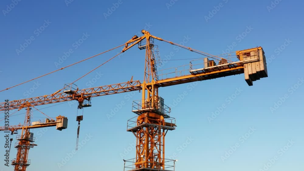 Yellow tower cranes on a construction site, against clear blue sky, illuminated by late afternoon sun. Aerial footage. Yellow tower cranes on a construction site, against clear blue sky, illuminated by late afternoon sun. Aerial footage.