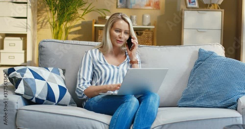 Pretty good looking woman talking on phone while searching internet on laptop on sofa in apartment. Beautiful Caucasian female chatting on phone with surprised face in morning room. Home concept photo