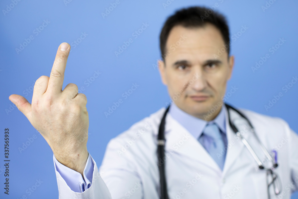 Middle-aged angry doctor in white medical coat and stethoscope showing middle finger doing fuck you, provocation and rude attitude, over blue background Middle-aged angry doctor in white medical coat and stethoscope showing middle finger doing fuck you, provocation and rude attitude, over blue background