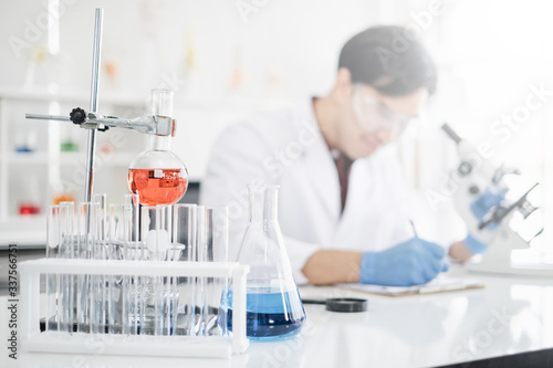 A male scientist with black hair wearing white coat and protective glassware writing and looking into a microscope in a laboratory setting with test tubes. Person blur in background.