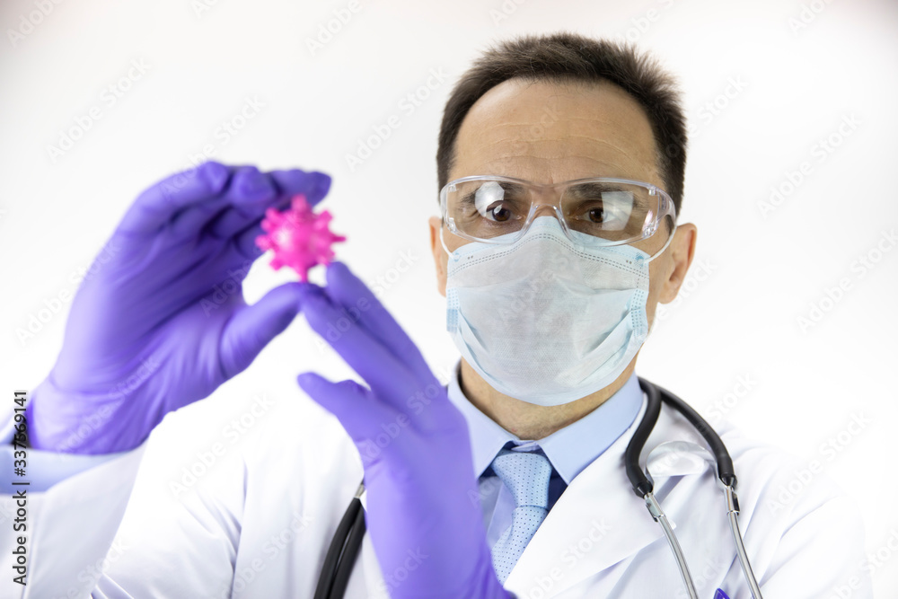 Male middle age lab technician assistant in medical uniform, protective mask and glasses holding covid-19 virus cell in hands. Close-up, focused on scientist. Coronavirus vaccine development Male middle age lab technician assistant in medical uniform, protective mask and glasses holding covid-19 virus cell in hands. Close-up, focused on scientist. Coronavirus vaccine development