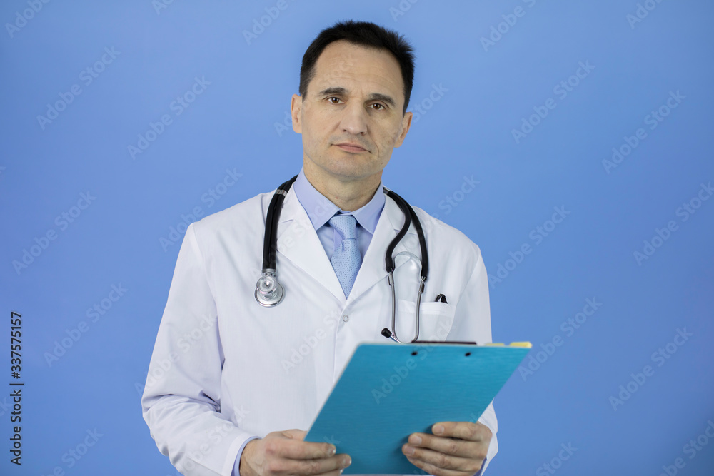 Portrait of a сaucasian adult doctor in a white coat with a stethoscope on his neck on a blue background. Serious stern look at the camera. Holding a blue folder with the results of patient analyzes. Portrait of a сaucasian adult doctor in a white coat with a stethoscope on his neck on a blue background. Serious stern look at the camera. Holding a blue folder with the results of patient analyzes.