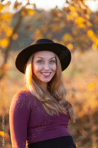Beautiful pregnant caucasian woman with long hair in hat outside in nature on warm sunny day. Smiles and looks at camera. Autumn nature backsides. Rest in park, meditation, retreat