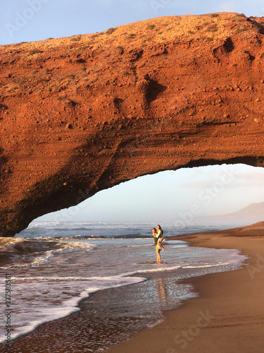 Couple in love walking under huge arch on Legzira ocean beach at sunset. Famous and popular landmark in Morocco. photo