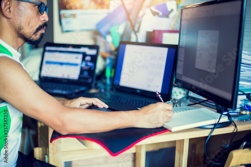 Asian man in eyewear working with computer remotely, sitting at wooden table in bed room. Pleasant serious man programming and testing, searching information online. The concept of work from home.