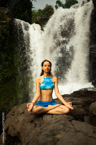 Young Caucasian woman sitting on the rock, meditating, practicing yoga at waterfall. Gyan mudra. Tegenungan waterfall in Ubud, Bali, Indonesia.