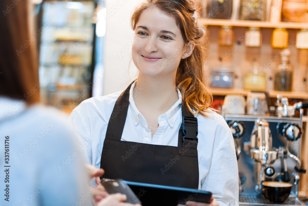 Young woman barista in brown apron is holding tablet, taking credit card from client in cafe. Smiling girl prepared americano espresso for customer. Professional coffee machine on background. Young woman barista in brown apron is holding tablet, taking credit card from client in cafe. Smiling girl prepared americano espresso for customer. Professional coffee machine on background.