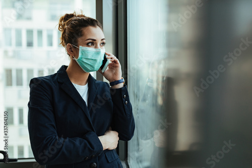Businesswoman with protective mask talking on mobile phone by the window.