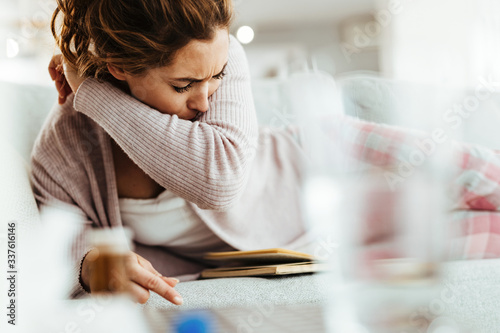 Sick woman sneezing into her elbow while lying down on sofa at home.