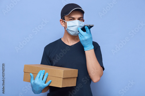 Studio shot of internet shop employee wearing protective mask and gloves to prevent spreading corona virus, courier sending voice message to his client, holding box with order in hand over blue wall. photo