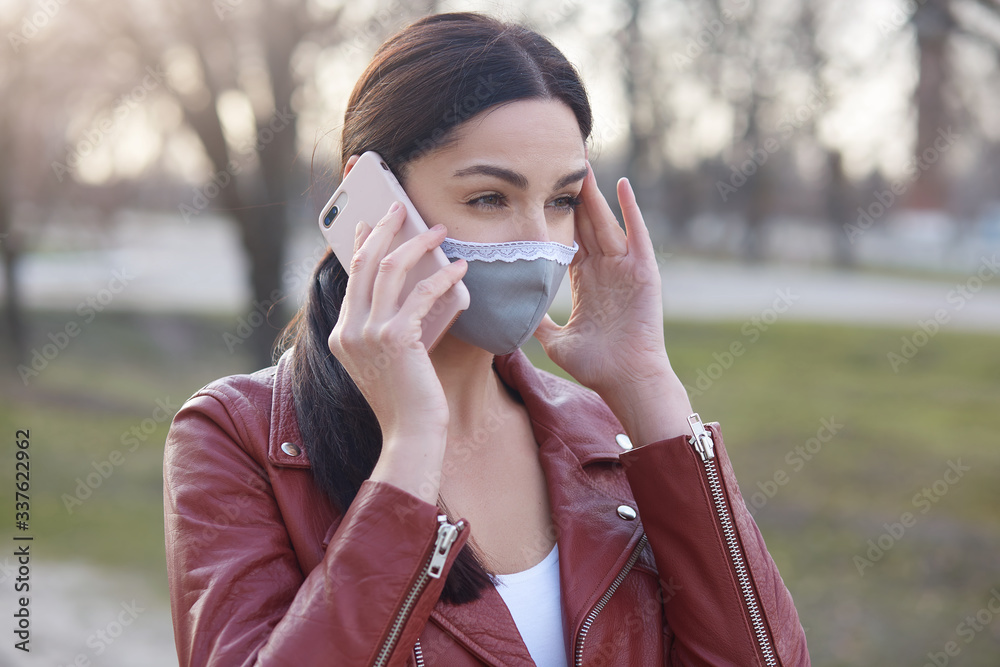 Closeup portrait of worried female wearing protective mask and stylish leather jacket, talking to phone with her grandmother, calls her on do not walk in open air, woman warns her family from Covid 19