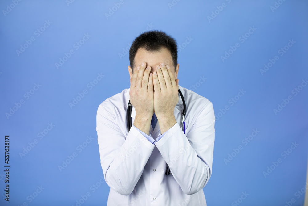Middle-aged handsome doctor in white coat and stethoscope shocked covering face with hands because of mistake from fatigue, blue background, fear concept Middle-aged handsome doctor in white coat and stethoscope shocked covering face with hands because of mistake from fatigue, blue background, fear concept