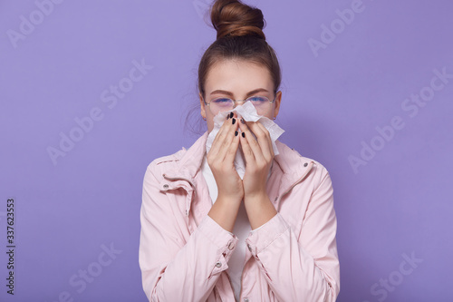 Fototapeta Naklejka Na Ścianę i Meble -  Horizontal shot of young female wearing pale pink jacket and glasses, has hair bun, looking at camera, using napkins for runny nose, posing isolated over lilac studio background. Healthcare concept.