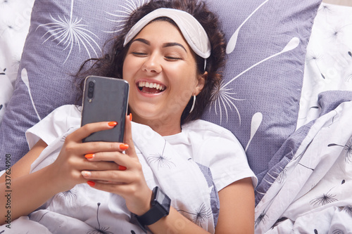 Image of attractive brunette girl lying on pillow with smart phone in hands, wearing t shirt and blindfold, female smiling happily, reading message from her boyfriend, having smart watch on her wrist photo