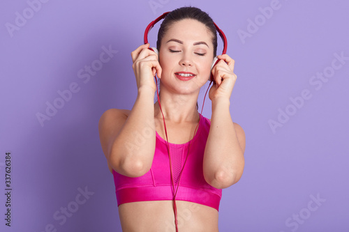 Image of sportswoman posing with closed eyes in studio, against lilac wall, female dresses sports wear, enjoying listening to music during workout, sporty girl has one minute rest between sets.