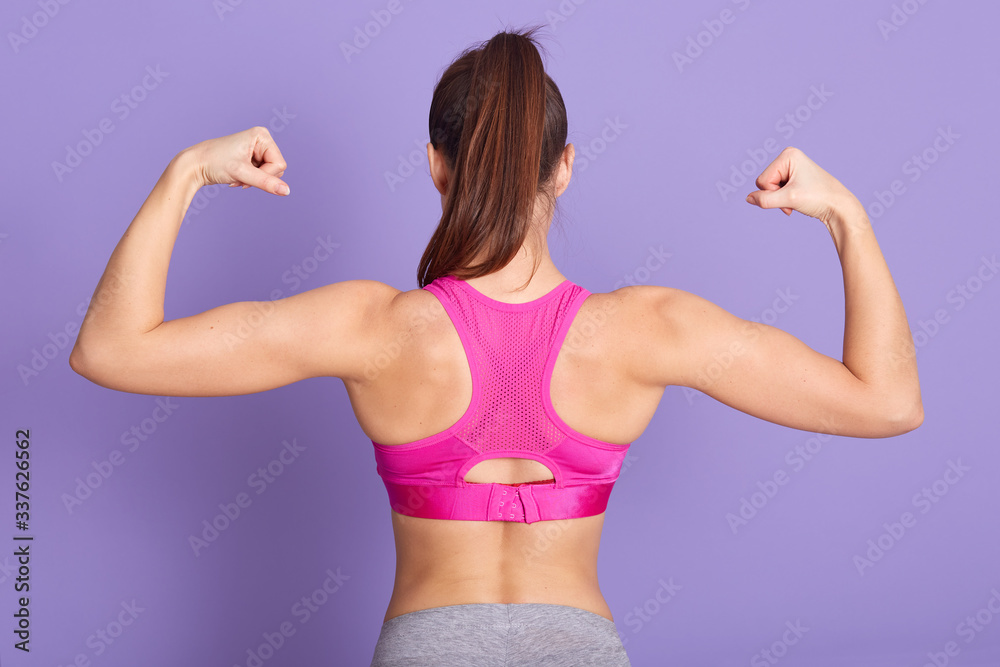 Studio shot of female posing backwards, wearing rosy top, standing against lilac studio wall and showing her muscles, girl training in gym during long time, having excellent results. Fitness concept. Studio shot of female posing backwards, wearing rosy top, standing against lilac studio wall and showing her muscles, girl training in gym during long time, having excellent results. Fitness concept.