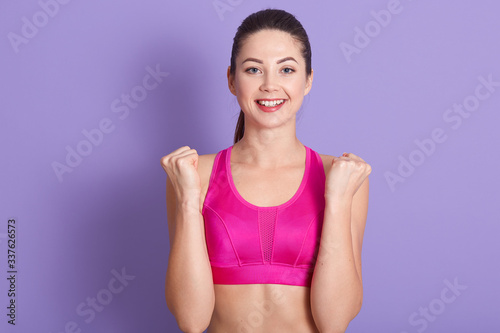 Closeup portrait of young attractive sportswoman wearing rose sports bra  standing isolated over lilac studio background and clenching fists  celebrating her success  great results. Fitness concept.