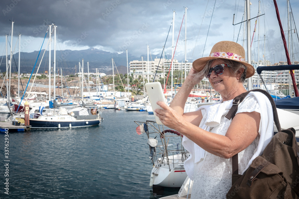 Senior attractive woman with backpack. A tablet in the hand. Boats and harbour on background