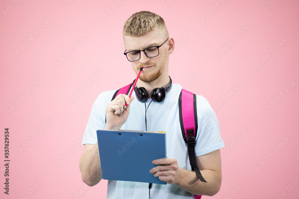 Portrait of thoughtful student in t-shirt and headphones over neck with notepad on hands, holds pen near mouth, thinking of solving a difficult task. Isolated pink background. Education concept. Portrait of thoughtful student in t-shirt and headphones over neck with notepad on hands, holds pen near mouth, thinking of solving a difficult task. Isolated pink background. Education concept.