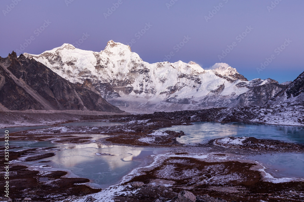 Highland icy wanderland. Mount Cho Oyu (8188m) basecamp in Himalayas, Nepal. High altitude lakes and mountain ridges in pinkish sunset light.