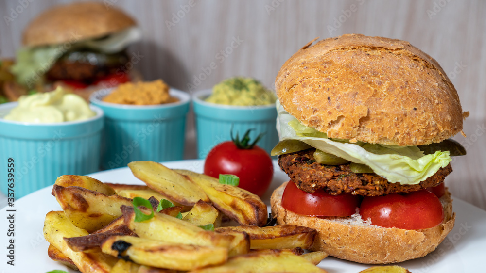 Vegan burger closeup with sliced tomatoes, pickled cucumbers, lettuce and baked potatoes. Homemade comfort plant based food. Cooking at home. Vegan burger closeup with sliced tomatoes, pickled cucumbers, lettuce and baked potatoes. Homemade comfort plant based food. Cooking at home.